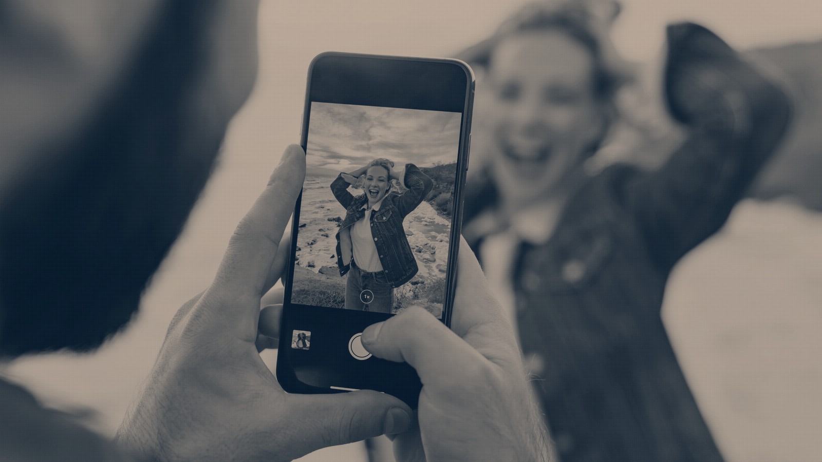 A couple on a clifftop at sunset, one photographing the other on a phone whose screen frames her mid-laugh, in a navy duotone